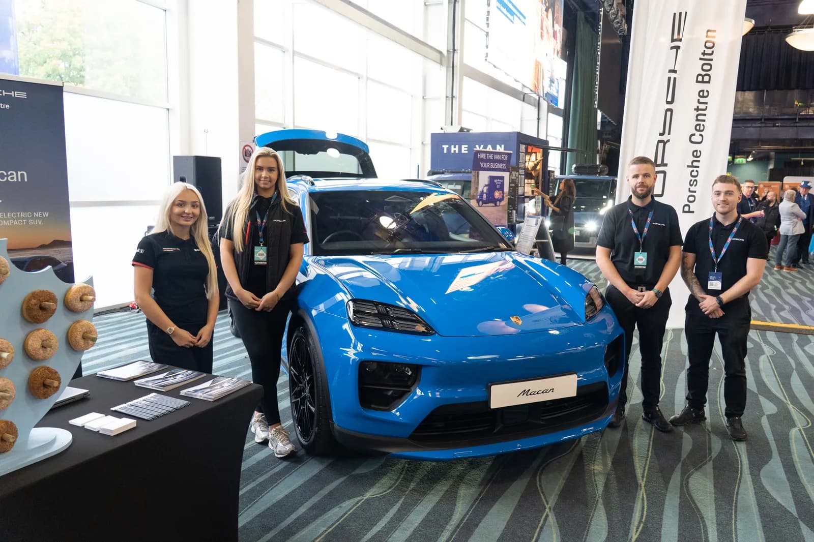 A group of five sales representatives stand around a blue Porsche Macan, with a display for Porsche Centre Bolton and a donut stand in the foreground.